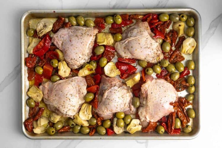 six chicken thighs arranged on a sheet pan, surrounded by olives, artichokes, and roasted red pepper strips.