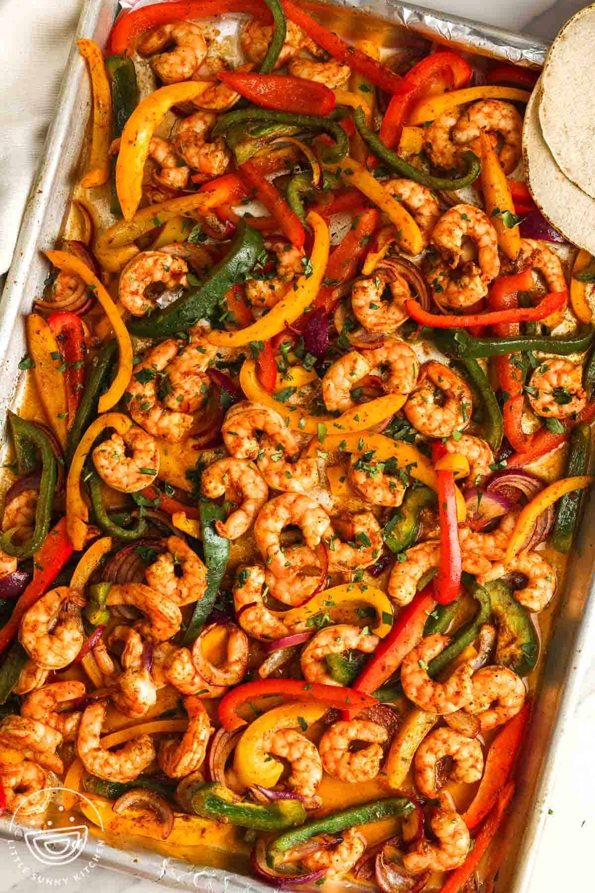 overhead view of a sheet pan meal of shrimp fajitas with veggies. With the shrimp and peppers are two white corn tortillas.