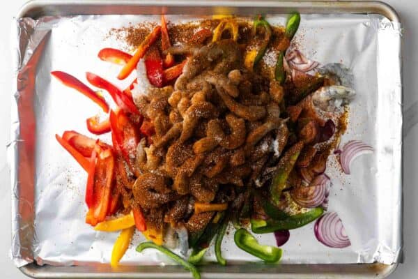 Seasoned raw shrimp, red, yellow, and green slices of bell pepper, and sliced red onion on a foil lined sheet pan.