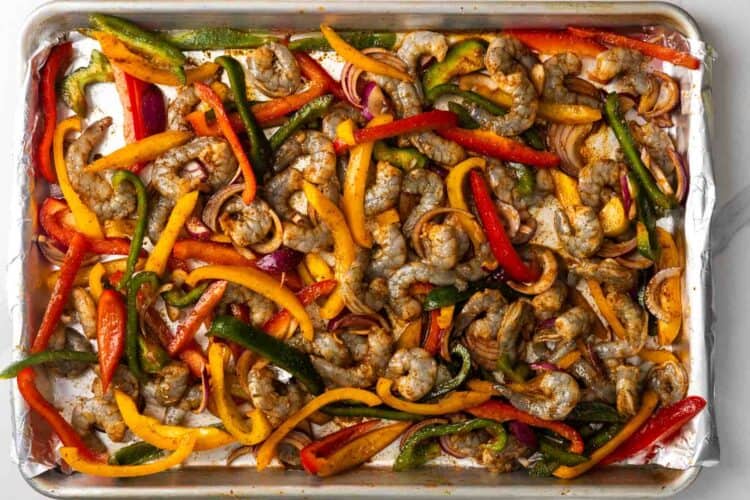 Shrimp, peppers, and onions arranged on a foil lined sheet pan, seasoned with fajita seasonings.
