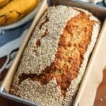 Tahini banana bread with sesame seed topping, baked in a rectangular loaf pan with brown parchment paper on the edges. Next to the loaf are two ripe bananas resting on a blue and white tea towel.