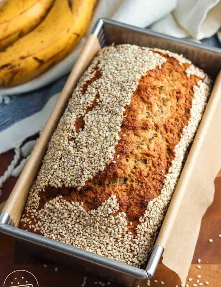 Tahini banana bread with sesame seed topping, baked in a rectangular loaf pan with brown parchment paper on the edges. Next to the loaf are two ripe bananas resting on a blue and white tea towel.