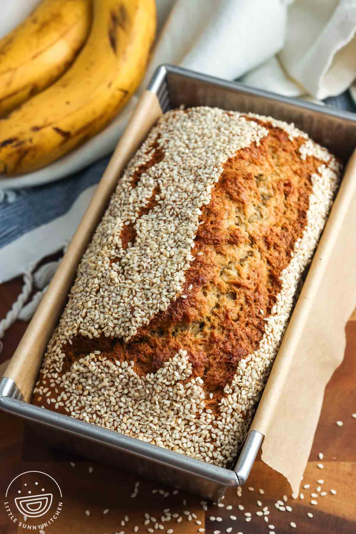 Tahini banana bread with sesame seed topping, baked in a rectangular loaf pan with brown parchment paper on the edges. Next to the loaf are two ripe bananas resting on a blue and white tea towel.