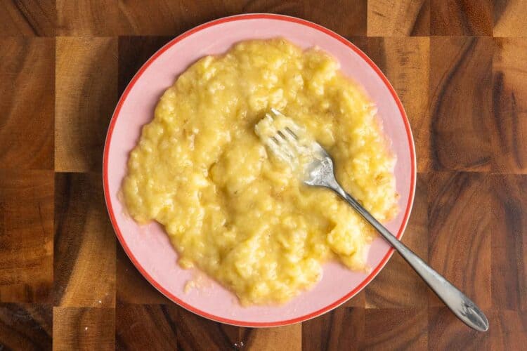 mashed banana on a pick plate with a fork. The plate is set on a wooden cutting board.
