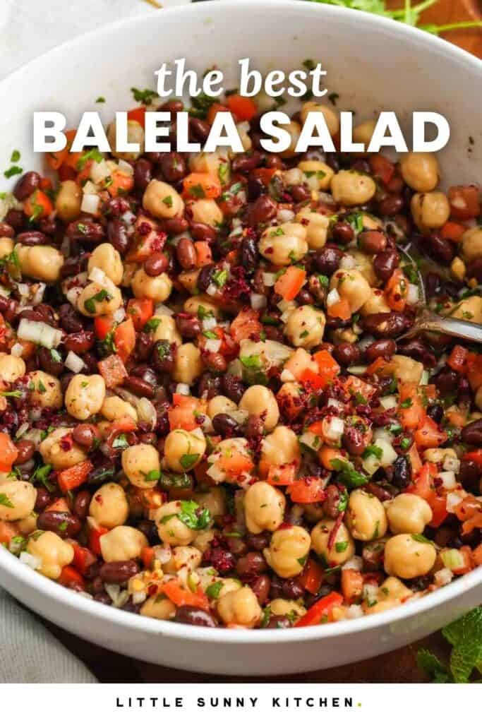 White bowl of balela salad with chickpeas, black beans, diced tomatoes, onion, fresh herbs, and sumac in a lemon olive oil dressing.