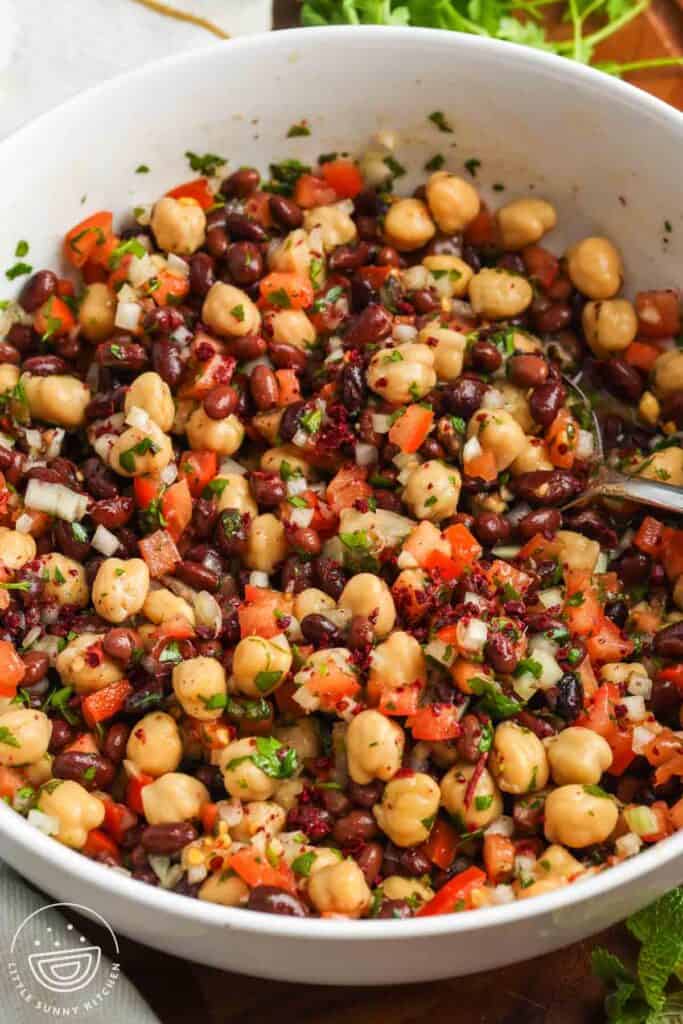 Close up of balela salad in a white bowl made with chickpeas, black beans, diced tomatoes, finely chopped onion, fresh herbs, and sumac tossed in a lemon and olive oil dressing.