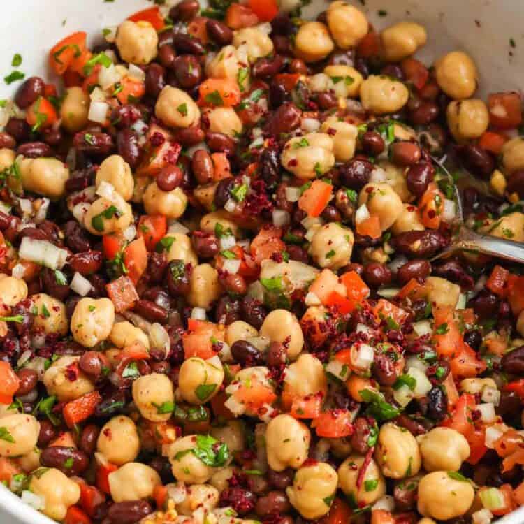Close up of balela salad in a white bowl made with chickpeas, black beans, diced tomatoes, finely chopped onion, fresh herbs, and sumac tossed in a lemon and olive oil dressing.