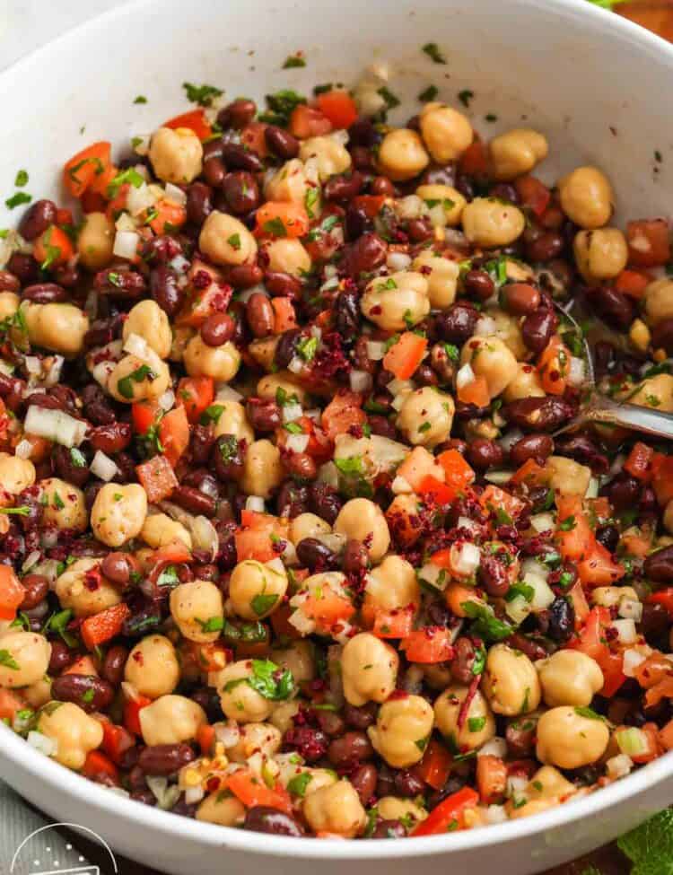 Close up of balela salad in a white bowl made with chickpeas, black beans, diced tomatoes, finely chopped onion, fresh herbs, and sumac tossed in a lemon and olive oil dressing.