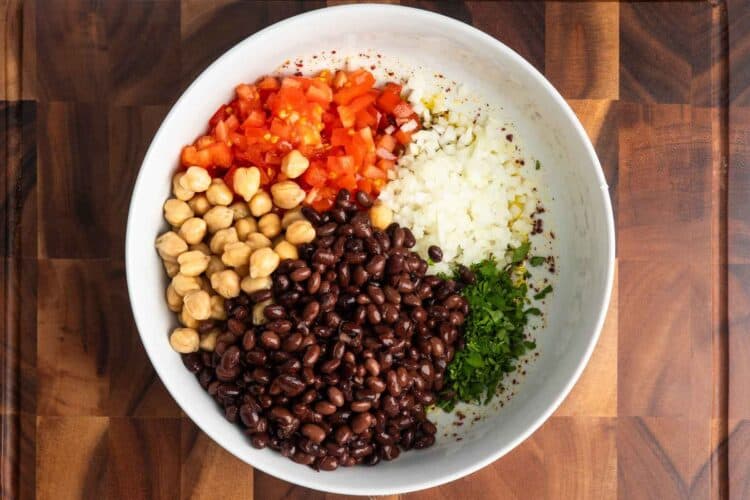 Chickpeas, black beans, diced tomatoes, finely diced yellow onion, and chopped fresh herbs added to the bowl with the dressing before mixing.