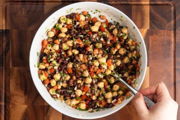 Balela salad fully mixed in a white bowl with chickpeas, black beans, tomatoes, onion, and herbs evenly coated in lemon and olive oil dressing.