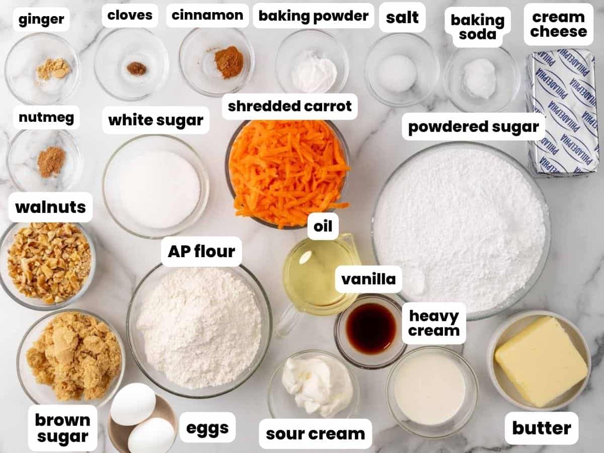 ingredients for carrot cake cupcakes, with walnuts and cream cheese for frosting. All of the cake ingredients are measured into small bowls and arranged on a marble surface.
