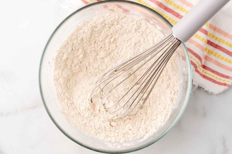 flour sifted with a whisk in a small glass bowl, next to a yellow and pink striped towel.