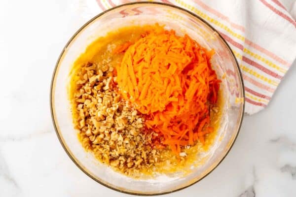 chopped nuts and grated carrots added to cupcake batter in a glass mixing bowl.