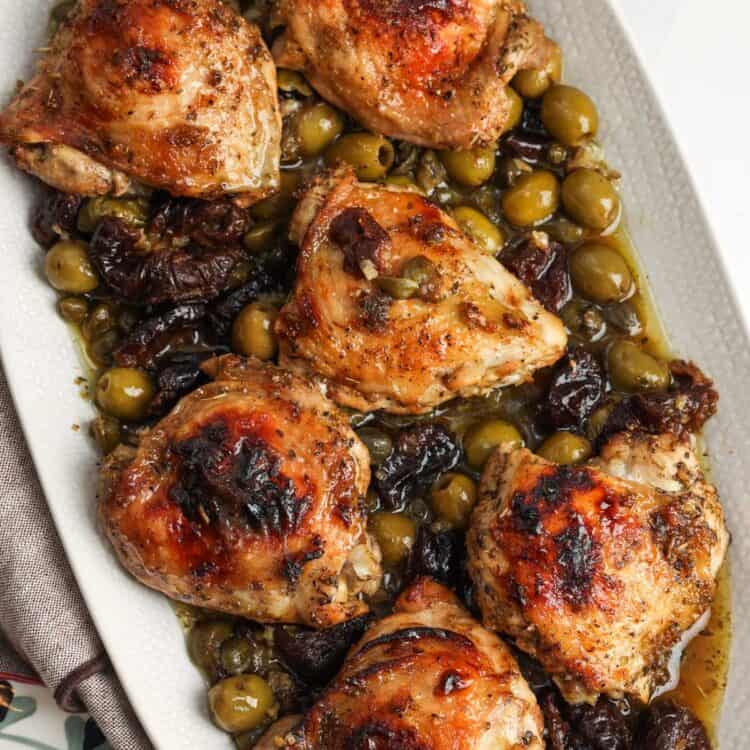 A white platter of chicken marbella, roasted chicken thighs marinated in olives, capers, prunes, and white wine.