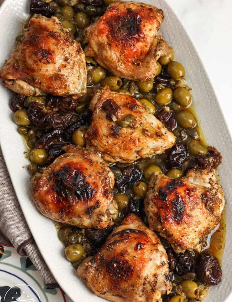 A white platter of chicken marbella, roasted chicken thighs marinated in olives, capers, prunes, and white wine.