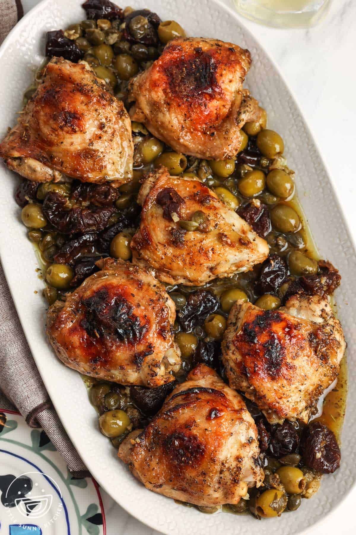 A white platter of chicken marbella, roasted chicken thighs marinated in olives, capers, prunes, and white wine.
