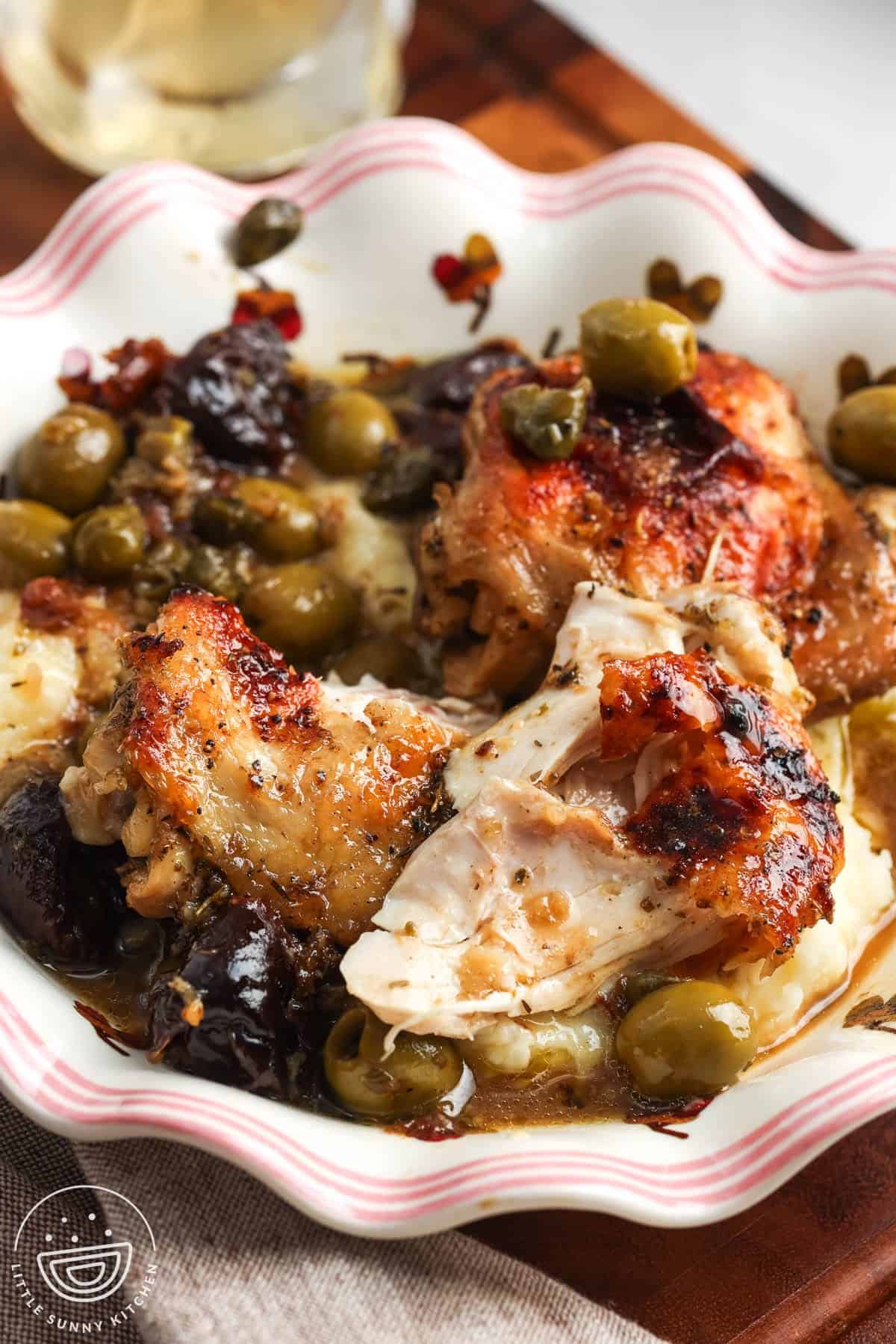 A fluted, decorative bowl of roasted chicken marbella with olives and prunes.