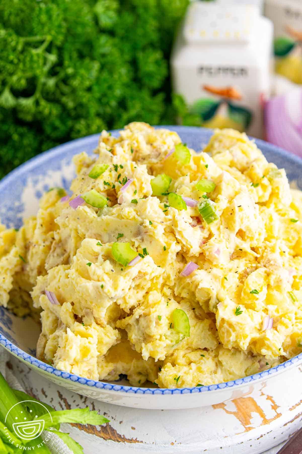 Close up of creamy smashed potato salad in a blue serving bowl, made with tender gold potatoes, mayonnaise dressing, celery, and red onion, with fresh parsley sprinkled on top.