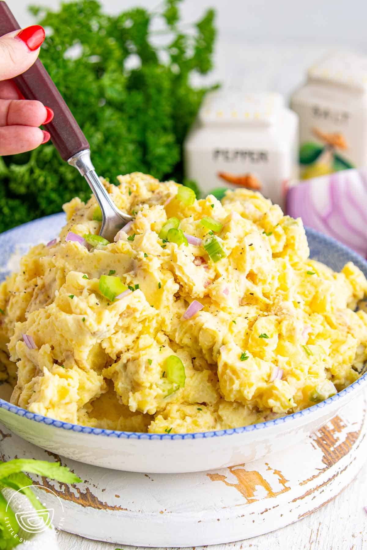 Hand holding a fork over a bowl of creamy smashed potato salad made with tender gold potatoes, mayonnaise dressing, chopped celery, red onion, and parsley.