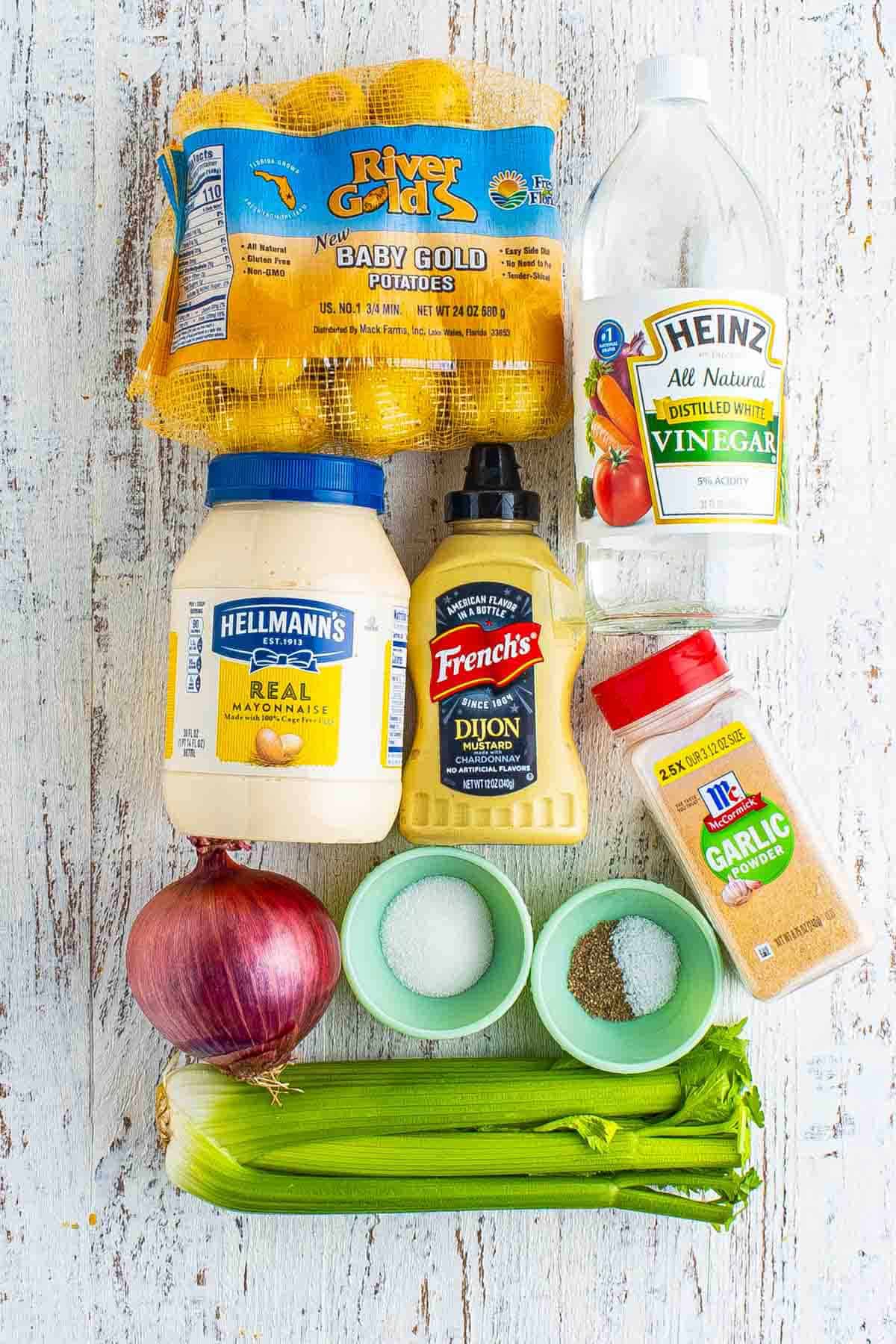 Ingredients for creamy smashed potato salad including baby gold potatoes, mayonnaise, Dijon mustard, white vinegar, garlic powder, red onion, celery, sugar, salt, and ground black pepper arranged on a white wooden surface.