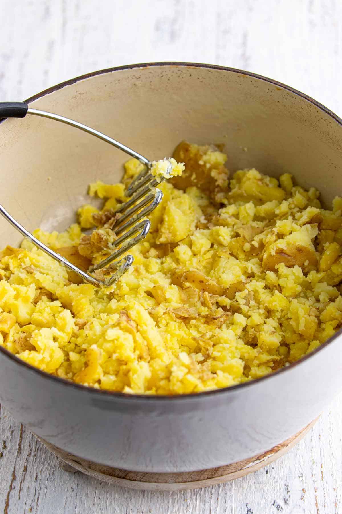 Cooked baby gold potatoes being gently mashed in a pot with a potato masher for creamy smashed potato salad.