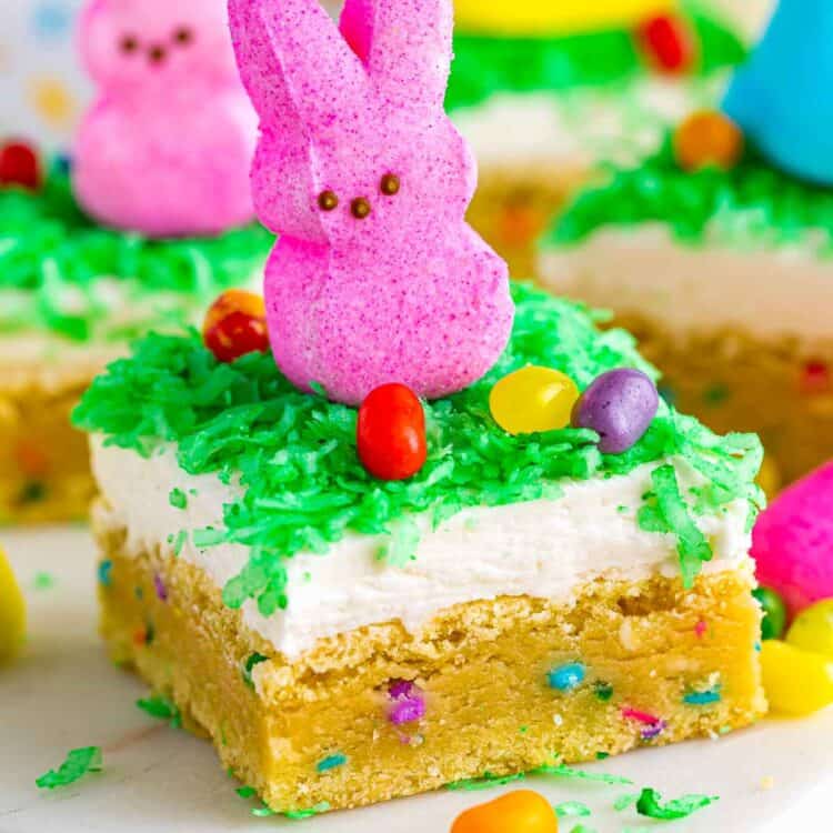 Easter sugar cookie bar topped with coconut frosting, green dyed coconut, a pink Peeps bunny, and colorful jelly beans on a soft sprinkle filled sugar cookie base.