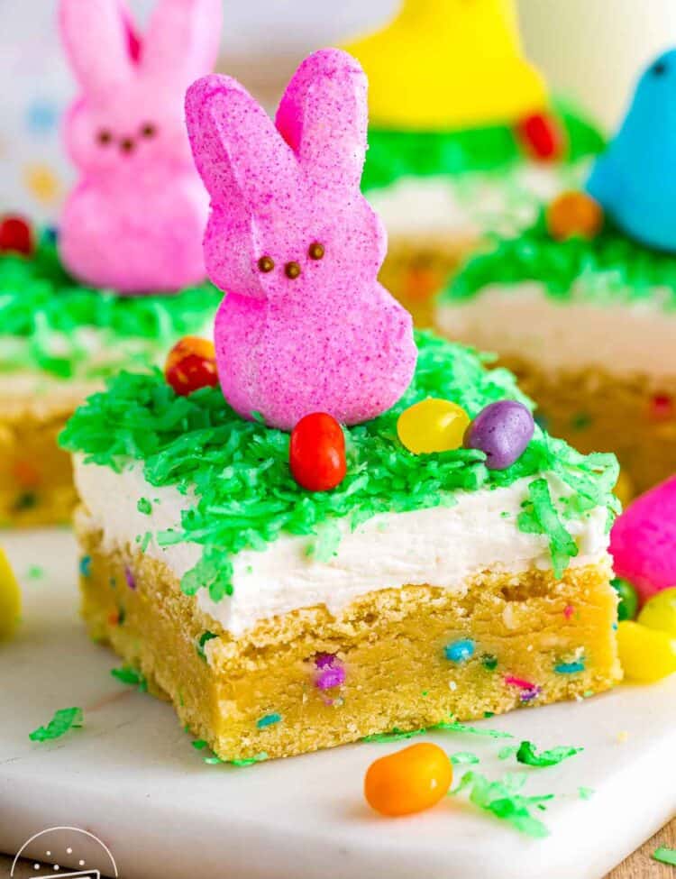 Easter sugar cookie bar topped with coconut frosting, green dyed coconut, a pink Peeps bunny, and colorful jelly beans on a soft sprinkle filled sugar cookie base.