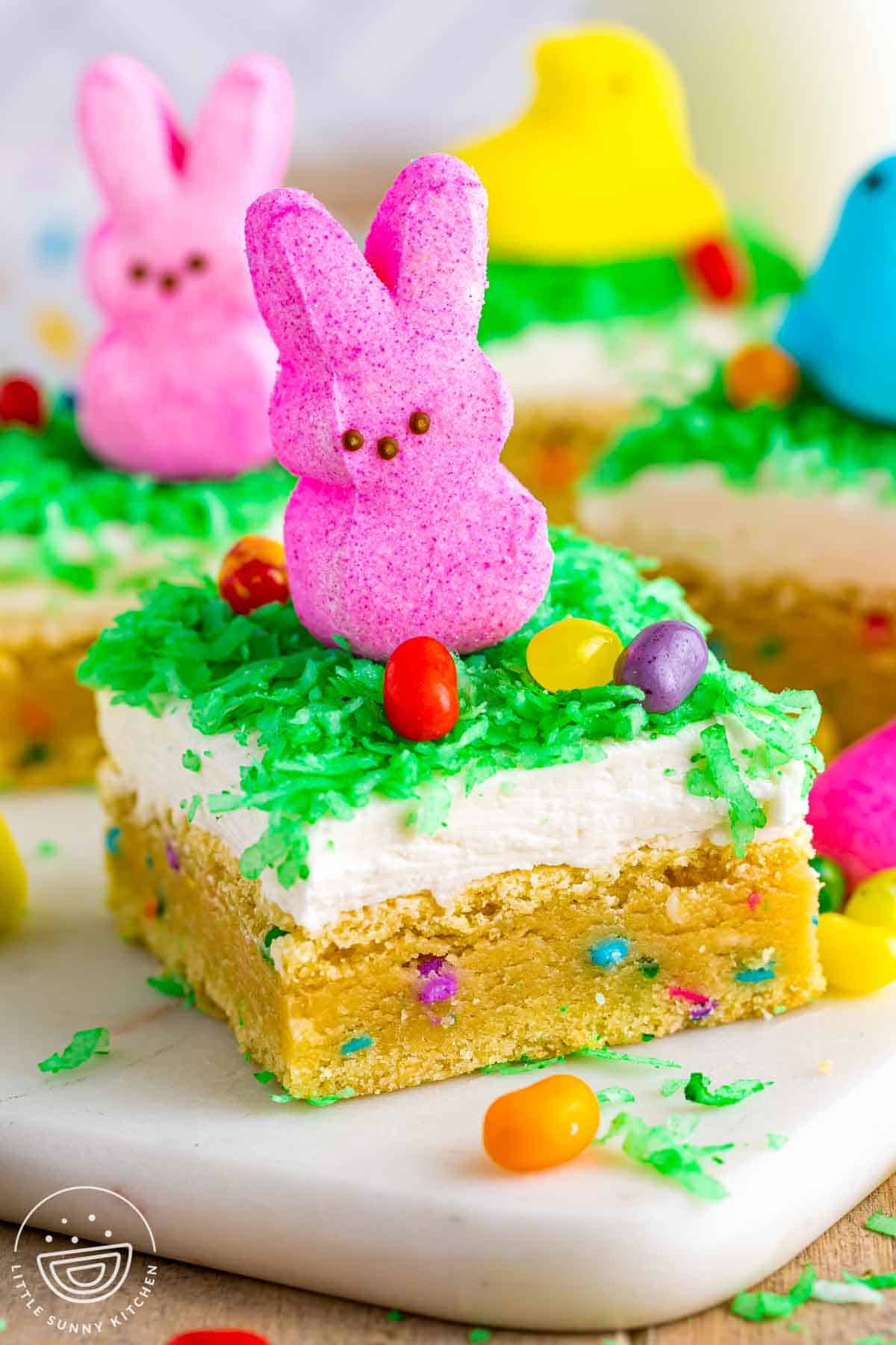 Easter sugar cookie bar topped with coconut frosting, green dyed coconut, a pink Peeps bunny, and colorful jelly beans on a soft sprinkle filled sugar cookie base.