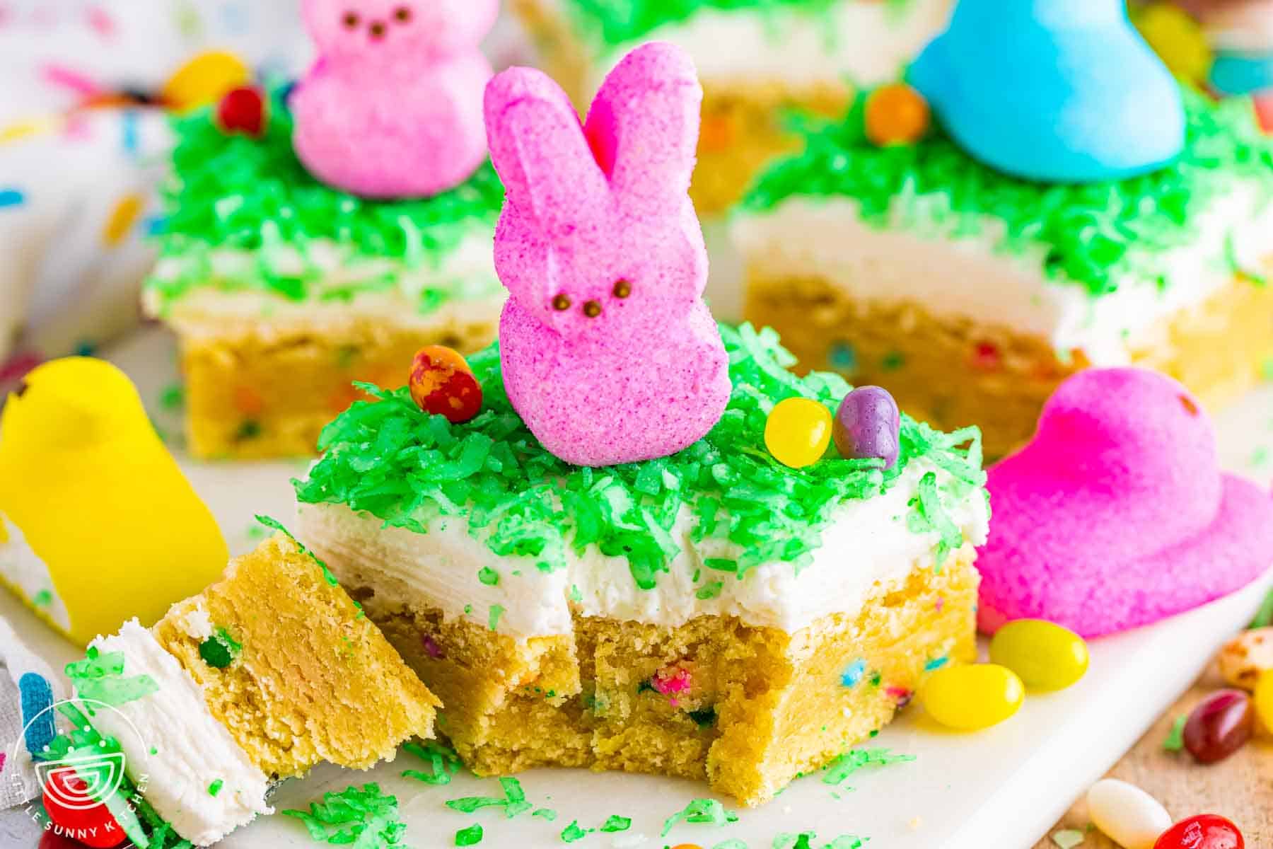 Easter sugar cookie bars topped with coconut frosting, green coconut flakes, a pink Peeps bunny, and colorful jelly beans, showing the soft sprinkle filled cookie base.