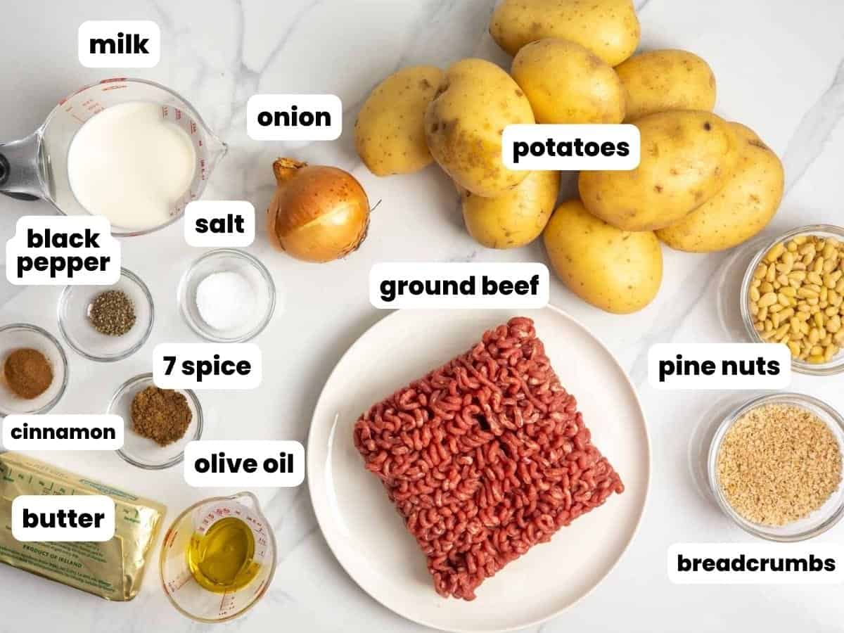 Overhead view of ingredients for Lebanese potato souffle including potatoes, ground beef, onion, milk, butter, olive oil, pine nuts, bread crumbs, salt, black pepper, seven spice, and cinnamon, each labeled on a marble surface.