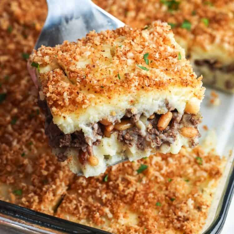 A spatula lifting a square slice of Lebanese potato souffle with layers of mashed potatoes and spiced ground beef, topped with golden buttered bread crumbs in a glass baking dish.