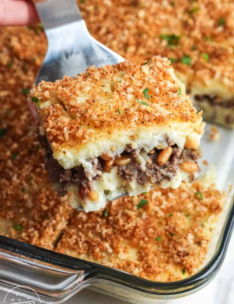 A spatula lifting a square slice of Lebanese potato souffle with layers of mashed potatoes and spiced ground beef, topped with golden buttered bread crumbs in a glass baking dish.