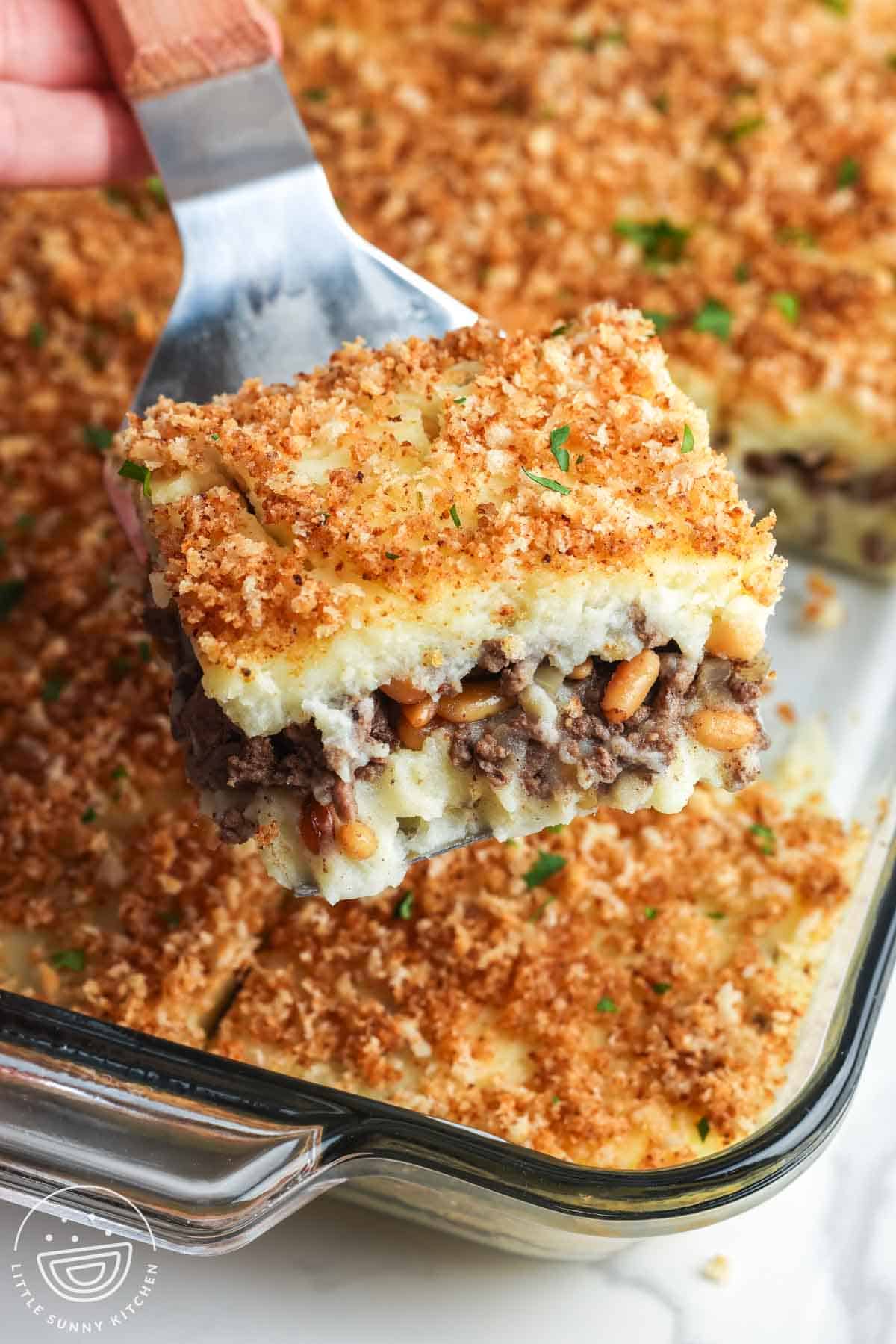 A spatula lifting a square slice of Lebanese potato souffle with layers of mashed potatoes and spiced ground beef, topped with golden buttered bread crumbs in a glass baking dish.