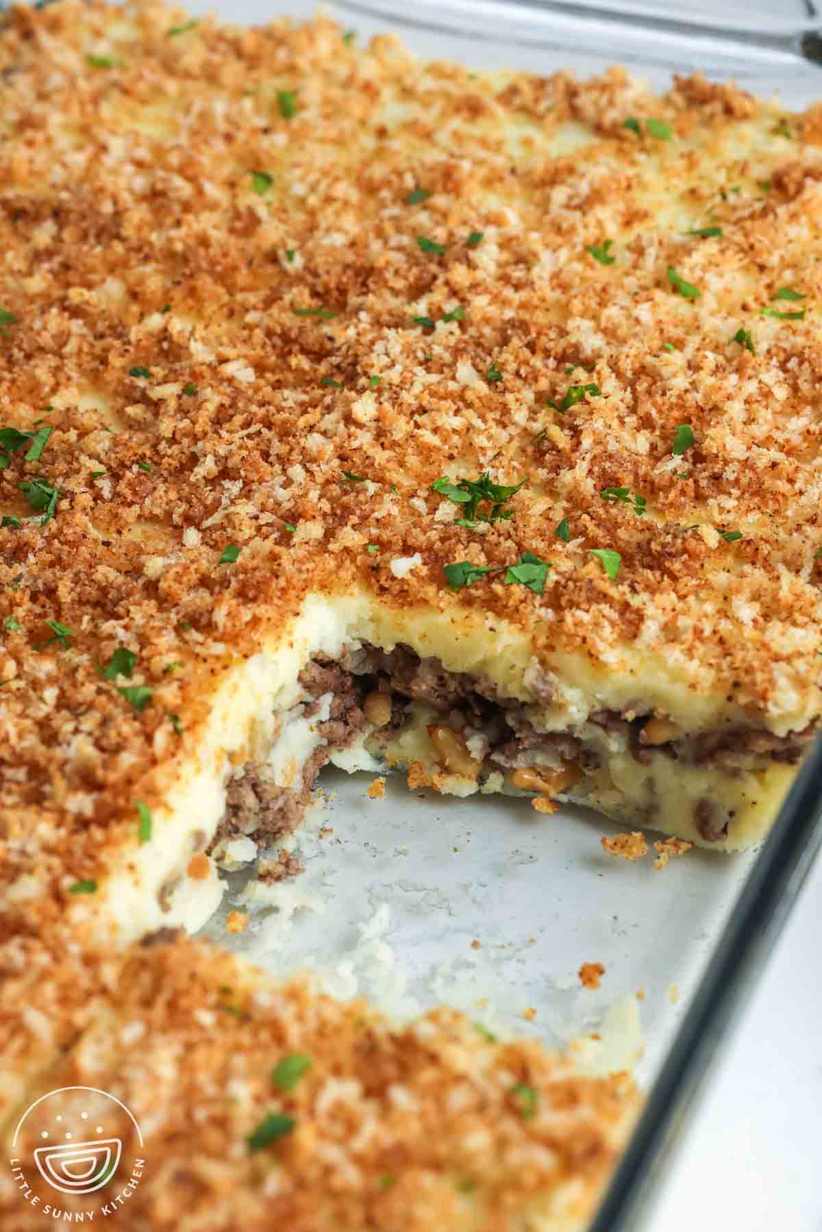 Lebanese potato souffle in a metal baking pan with a golden bread crumb topping, showing a cut out corner with layers of mashed potatoes and spiced ground beef.