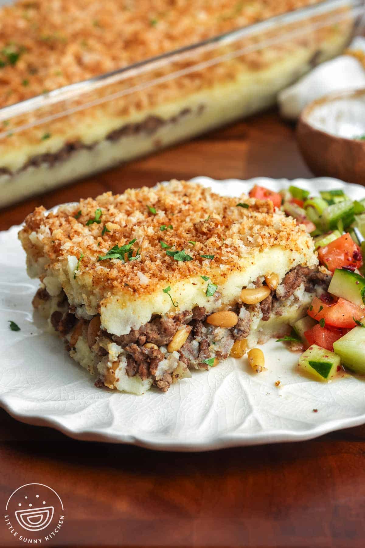 A slice of Lebanese potato souffle on a white plate with layers of mashed potatoes and spiced ground beef, topped with golden bread crumbs, served with chopped cucumber and tomato salad.