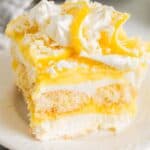 Lemon tiramisu slice with layers of mascarpone cream, lemon curd, and ladyfingers, topped with whipped cream, lemon slices, and white chocolate shavings.