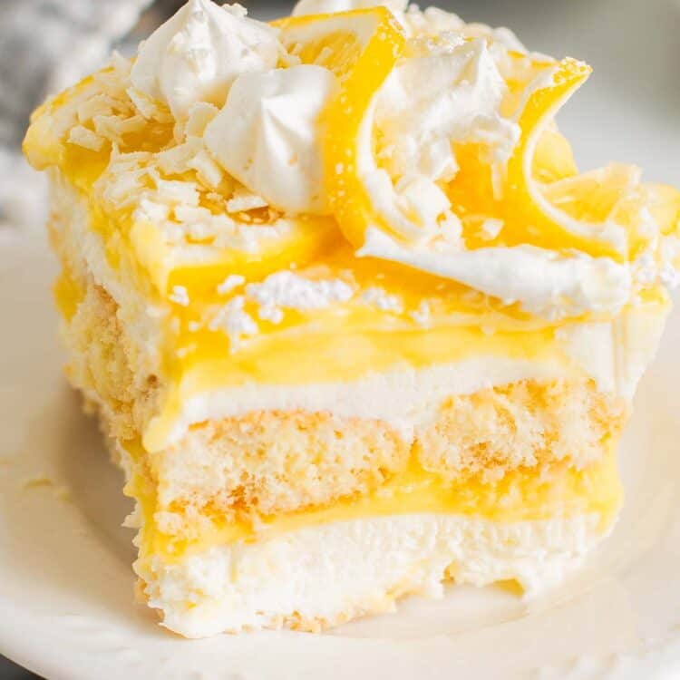 Lemon tiramisu slice with layers of mascarpone cream, lemon curd, and ladyfingers, topped with whipped cream, lemon slices, and white chocolate shavings.