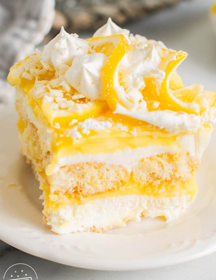 Lemon tiramisu slice with layers of mascarpone cream, lemon curd, and ladyfingers, topped with whipped cream, lemon slices, and white chocolate shavings.