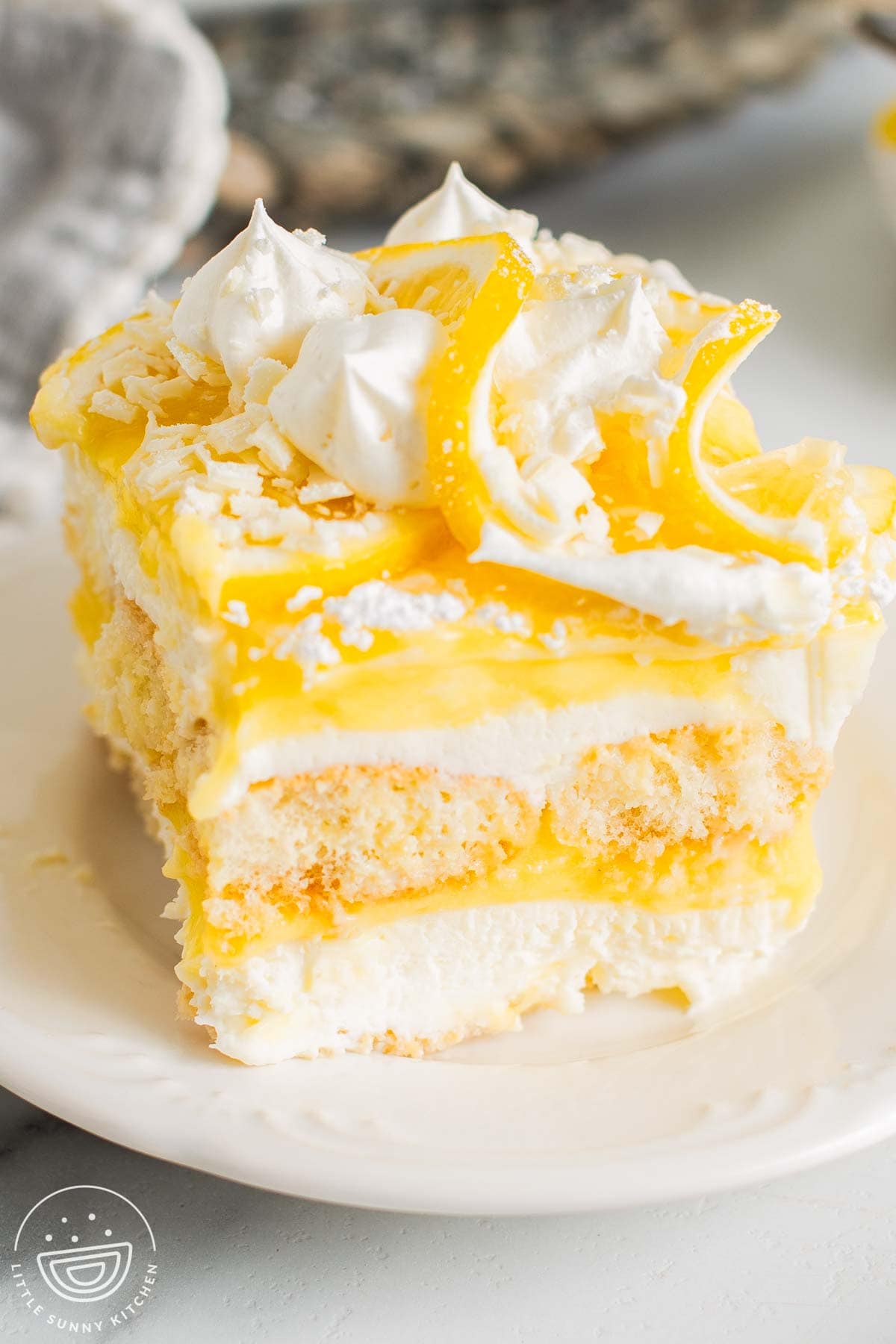 Lemon tiramisu slice with layers of mascarpone cream, lemon curd, and ladyfingers, topped with whipped cream, lemon slices, and white chocolate shavings.