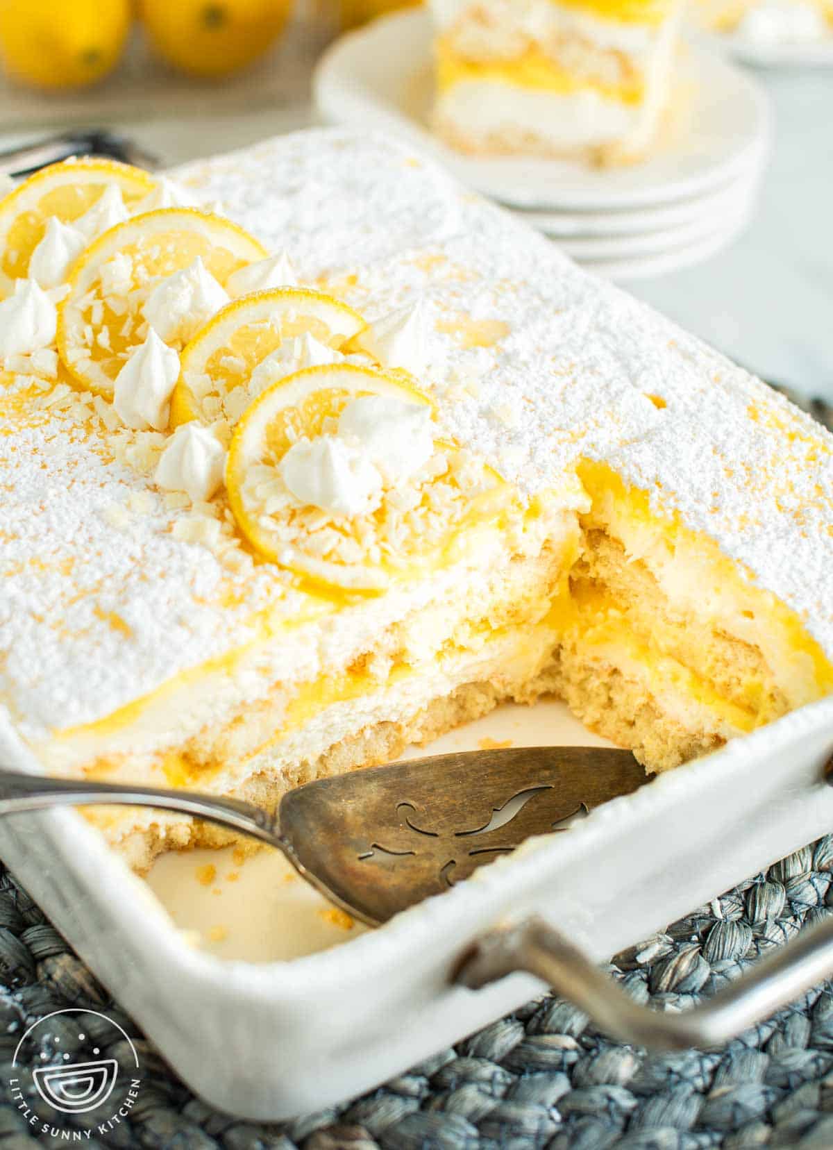 Lemon tiramisu in a baking dish dusted with powdered sugar and topped with lemon slices, whipped cream, and white chocolate, with a portion removed showing soft layered texture.
