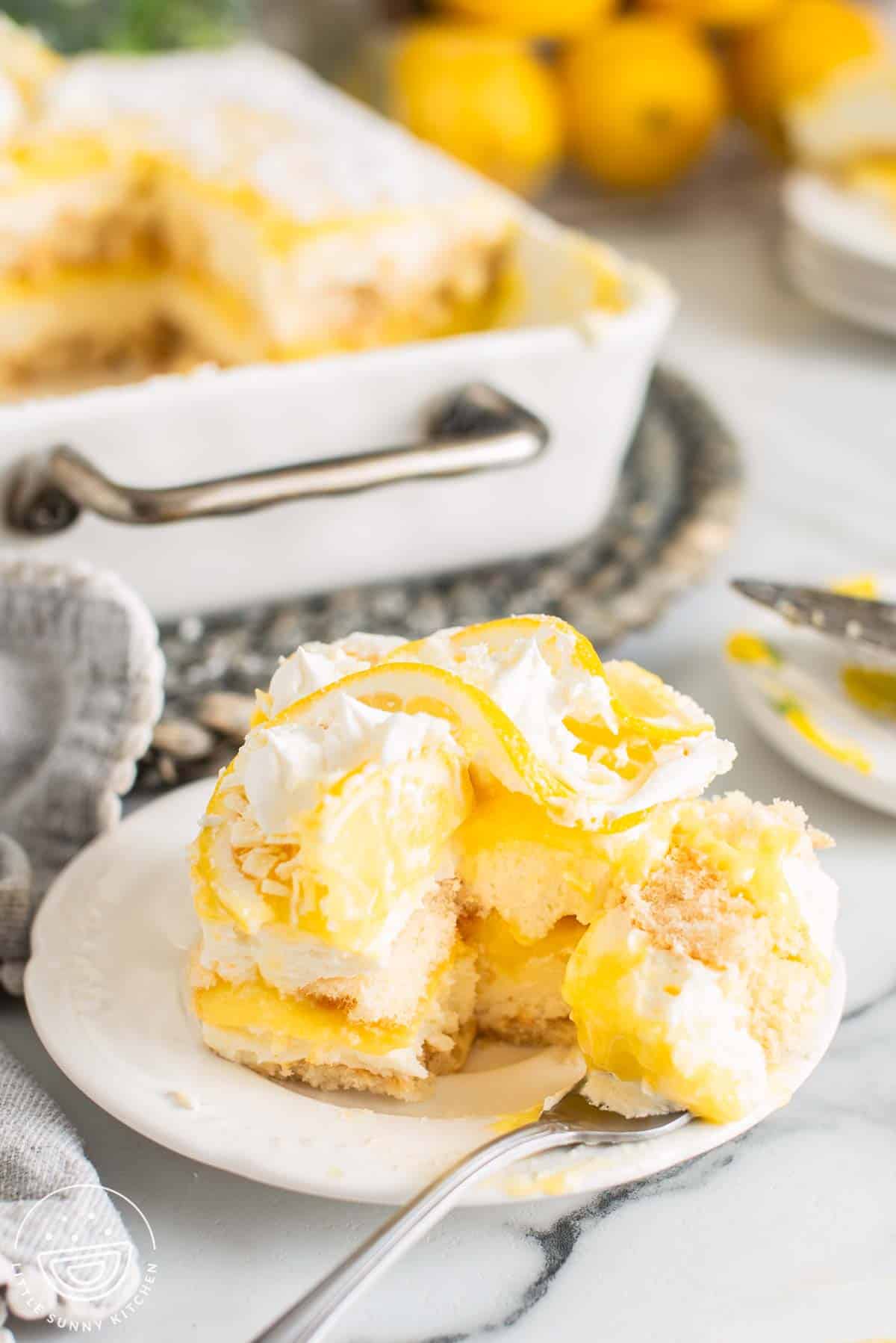 Lemon tiramisu served on a plate with a spoon, showing creamy mascarpone layers, lemon curd, and soft ladyfingers topped with whipped cream and lemon slices.