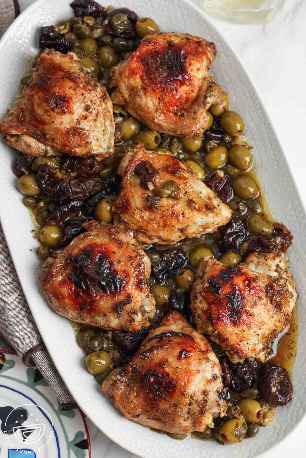 A white platter of chicken marbella, roasted chicken thighs marinated in olives, capers, prunes, and white wine.