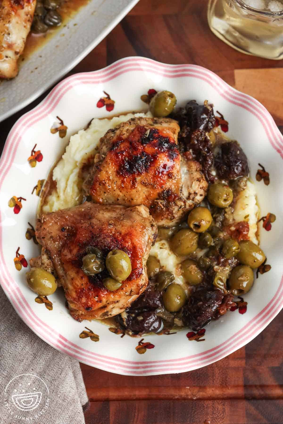 A pink edges bowl with an acorn design filled with mashed potatoes that are topped with two roasted chicken thighs, olives, capers, and prunes.