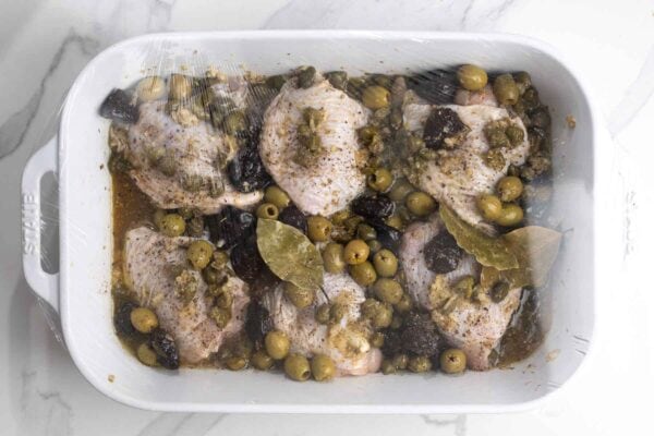 Raw chicken thighs marinating with olives, capers, prunes, seasonings, white wine, and bay leaves.