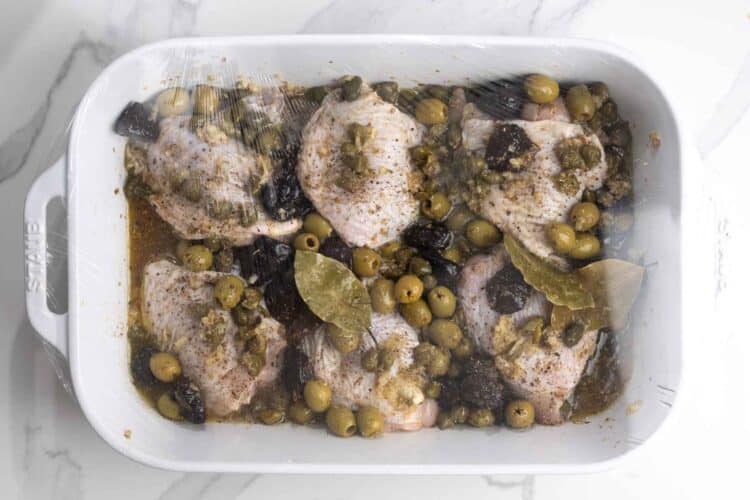 Raw chicken thighs marinating with olives, capers, prunes, seasonings, white wine, and bay leaves.