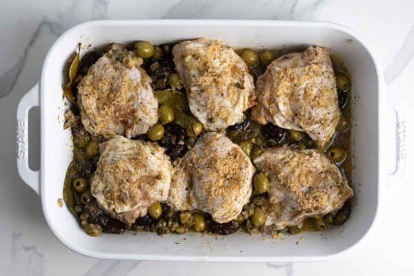 Seasoned raw chicken thighs on top of a bed of olives, capers, prunes, and bay leaves.