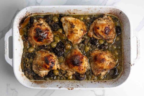 Chicken marbella baked in a white 9x13 -inch baking pan, includes six bone-in chicken thighs.