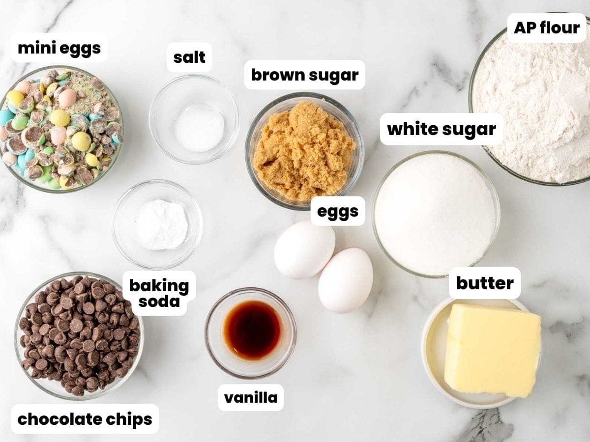 ingredients for making chocolate chip cookies with cadbury mini eggs. The eggs are crushed, and all of the ingredients are set out in bowls on a marble counter.