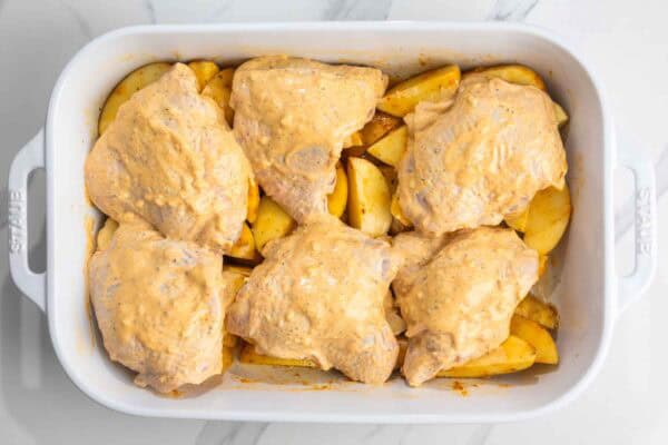 Chicken thighs coated in mayo and seasonings, set over seasoned raw potatoes in a roasting pan.
