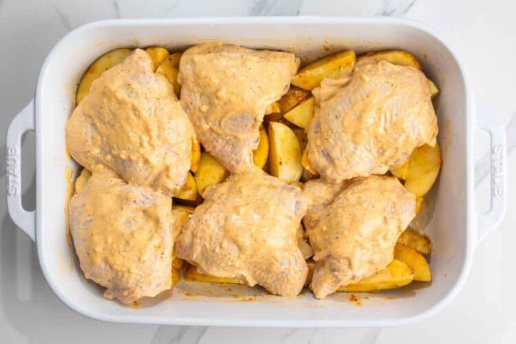 Chicken thighs coated in mayo and seasonings, set over seasoned raw potatoes in a roasting pan.